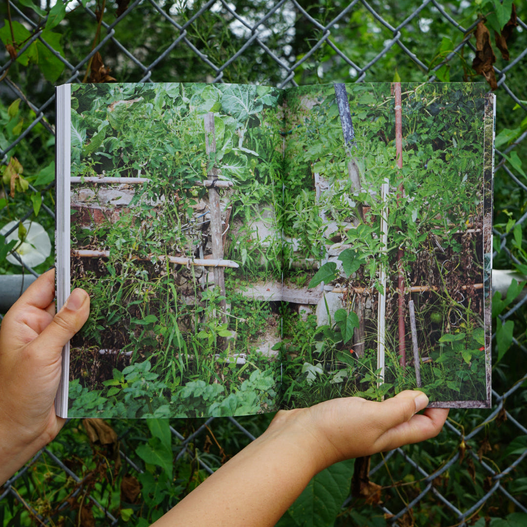 A person holds an open book showing a garden scene with green plants and a fence, photographed outdoors in front of a chain-link fence with greenery.