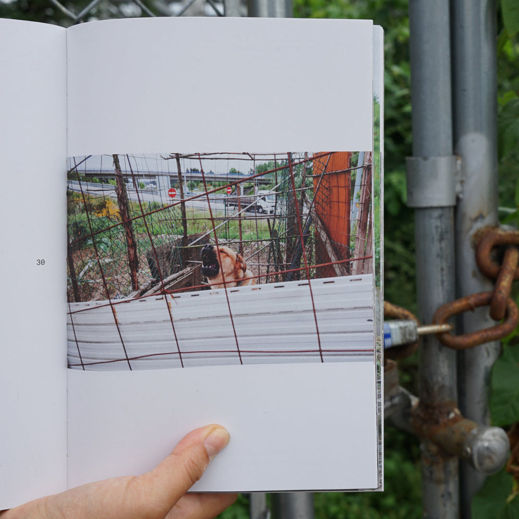 A hand holds an open book in front of a chain-link fence and metal gate. The open page shows a printed photograph depicting a barking dog behind wire mesh in a garden located next to a highway.