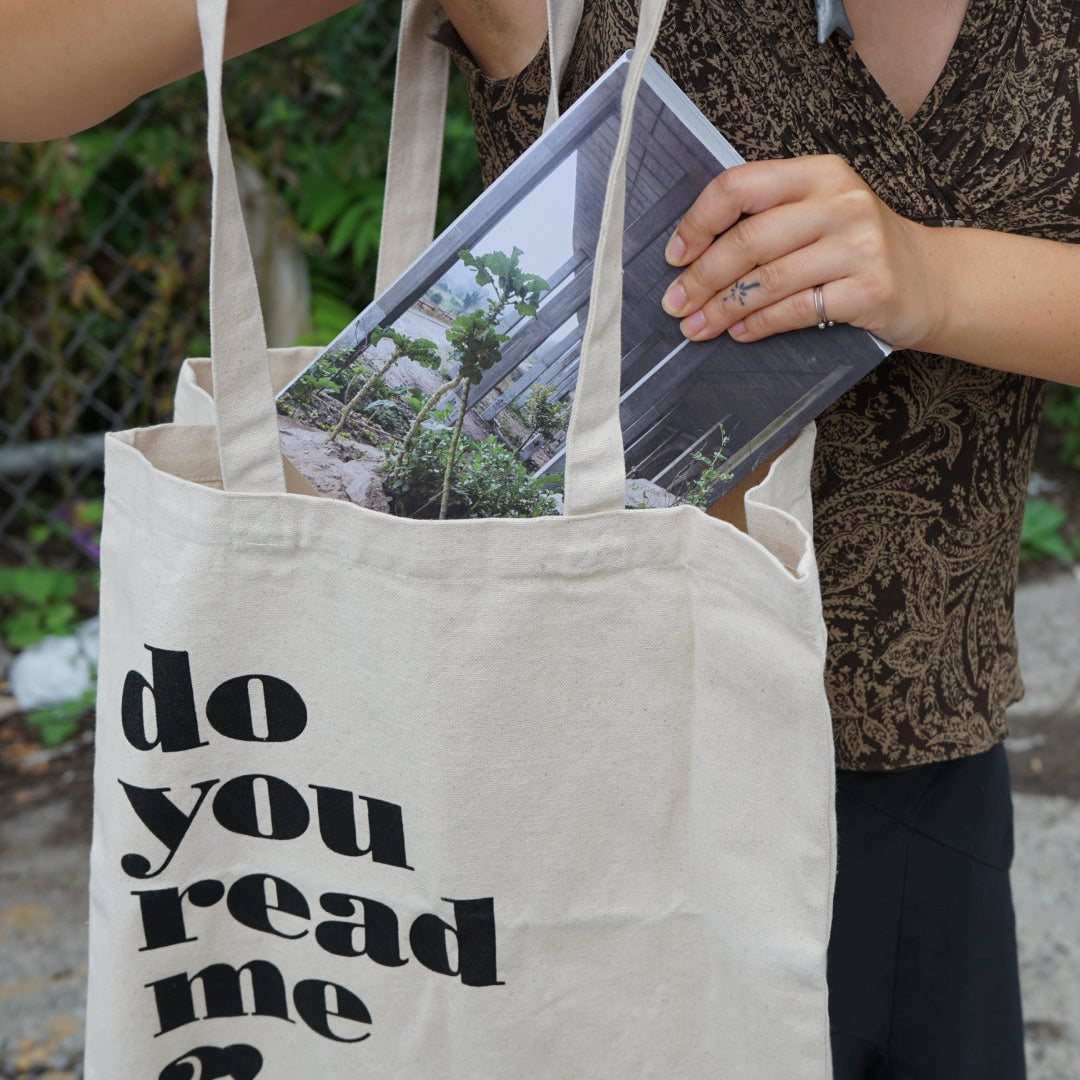 A person wearing a canvas tote bag with black text reading 'do you read me ?!' outdoors, pulls out a book from inside the tote.